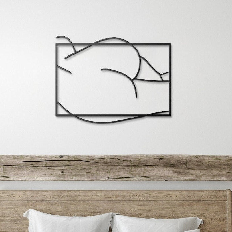 A minimalist black line art representation of a nude female figure on a metal wall sign, displayed above a bed in a room with a wooden headboard and decor.